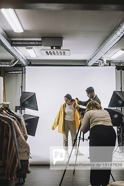 Photographer photographing designer helping model wearing jacket against white backdrop in studio