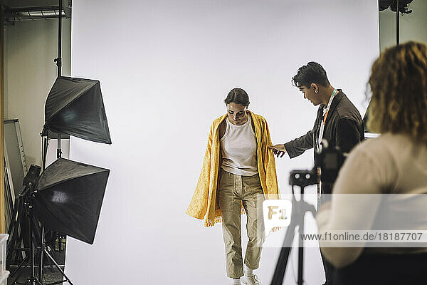 Male designer assisting female model against white backdrop during photo shoot in studio