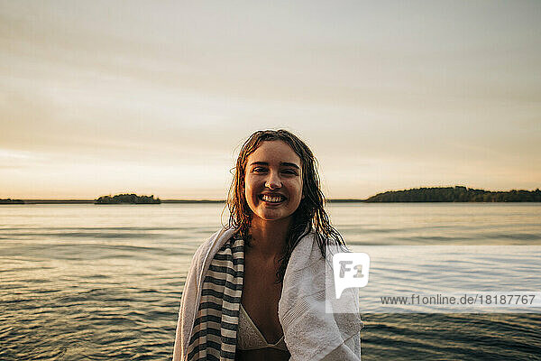 Portrait of happy woman with towel against lake during sunset