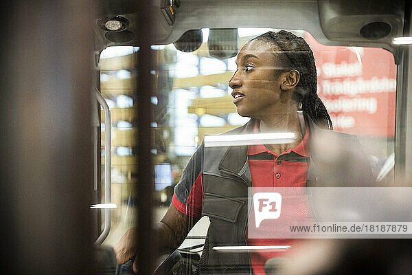 Female warehouse worker looking away while sitting in forklift seen through windshield