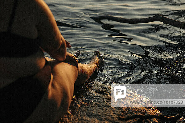 Low section of young voluptuous woman sitting by water during sunset