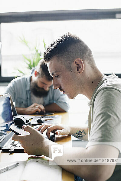 Männlicher Computerprogrammierer  der ein Smartphone neben einem Laptop auf einem Schreibtisch in einem Startup-Unternehmen benutzt