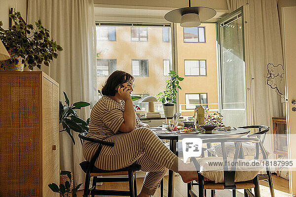 Junge Frau spricht mit ihrem Smartphone  während sie zu Hause auf einem Stuhl sitzt