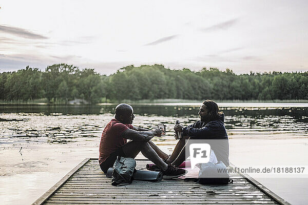 Man sharing smart phone with friend sitting on jetty at lake