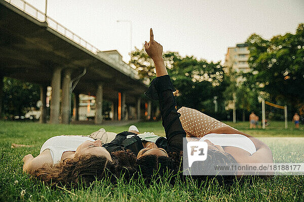 Teenager-Mädchen mit erhobener Hand  die auf einen Punkt zeigt  während sie inmitten von Freunden auf einer Wiese im Park liegt