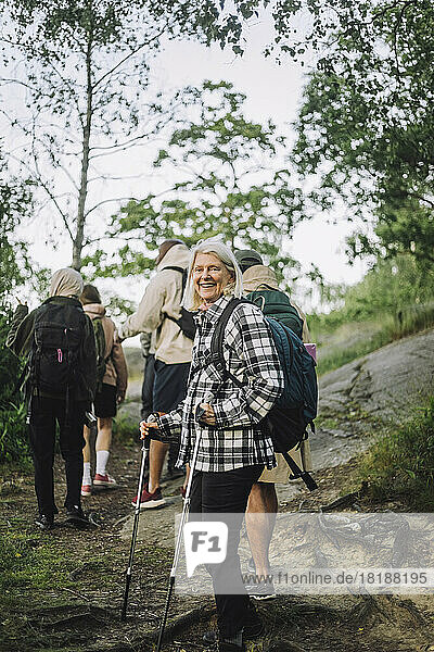 Seitenansicht einer lächelnden älteren Frau  die beim Wandern mit Freunden im Wald einen Stock hält