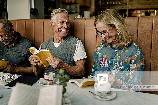 Glücklicher älterer Mann und Frau im Gespräch miteinander beim Lesen von Büchern durch einen männlichen Freund im Café