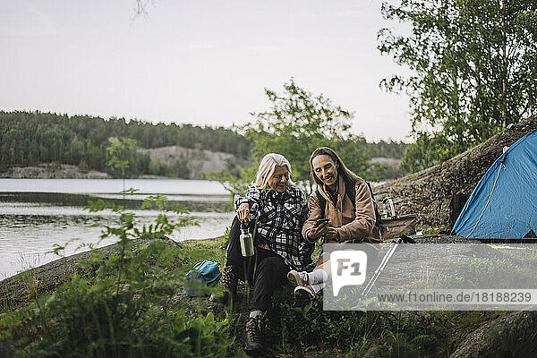 Ältere Freundinnen teilen ihr Smartphone  während sie beim Camping auf einem Felsen sitzen