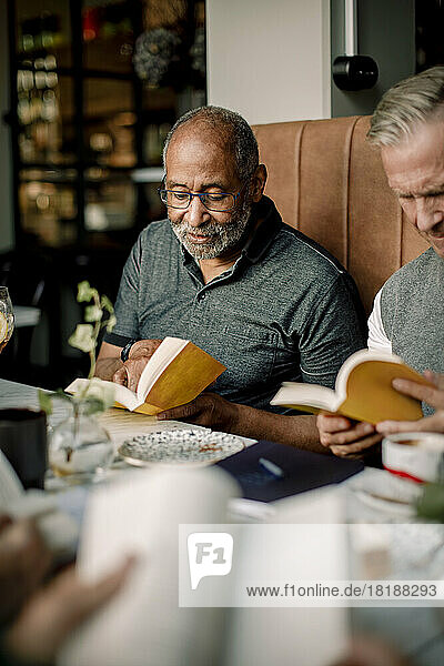 Senior man wearing eyeglasses reading book while sitting by male friend in cafe