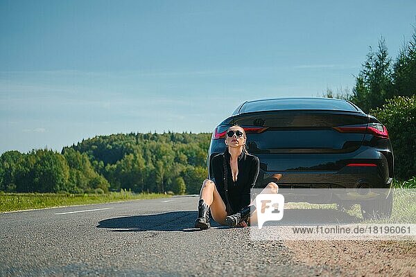 Strong and sexy woman in bodysuit and rough boots sits on asphalt by her powerful racing car
