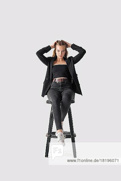 Porträt in voller Länge von schönen jungen Mädchen sitzen auf hohen Stuhl in schwarzen Jeans und Jacke über grauen Hintergrund