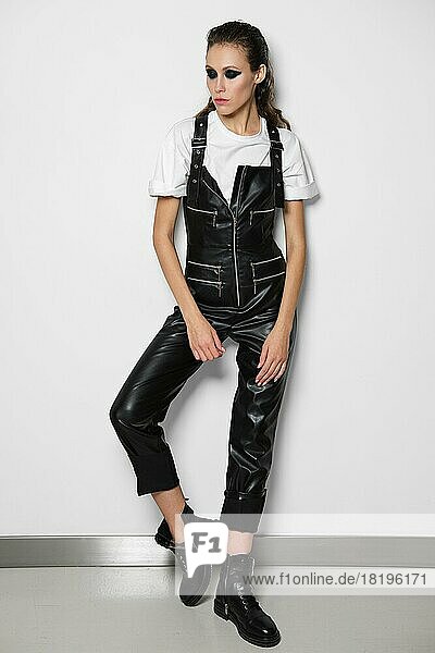 Girl in faux leather overalls and white t-shirt posing near white wall