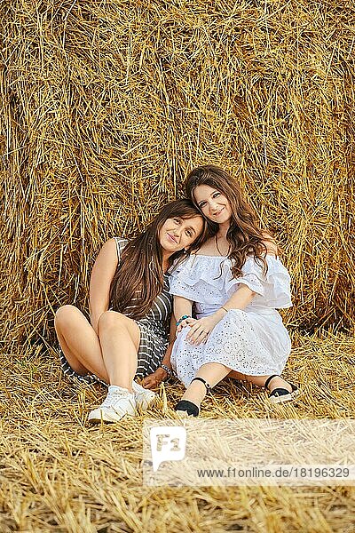 Two girlfriends are sitting leaning against a stack on a field