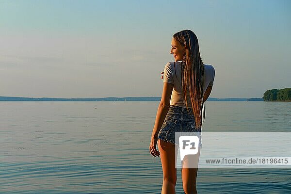 Hübsche Frau in Jeans-Shorts und weißem Unterhemd steht am Wasser in einem Erholungsgebiet bei Sonnenaufgang