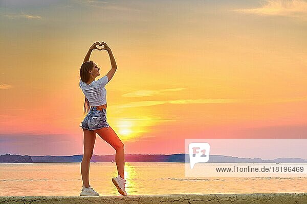 Junge Frau steht während des Sonnenuntergangs auf einem Pier und faltet ihre Handflächen in Form eines Herzens