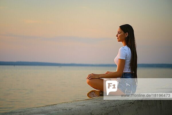 Seitenansicht einer Frau in Meditationshaltung auf dem Pier während des Sonnenuntergangs im Sommer
