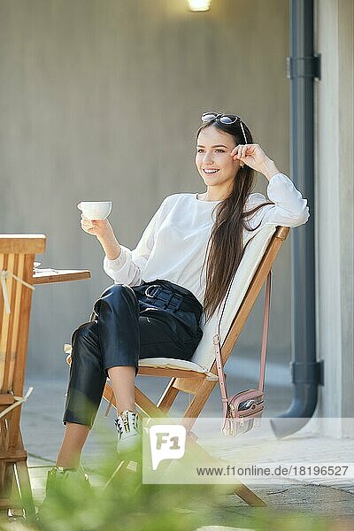 Happy young woman sitting on the terrace of the cafe in sunny day and drinking coffee. Woman enjoying first spring sun outdoor