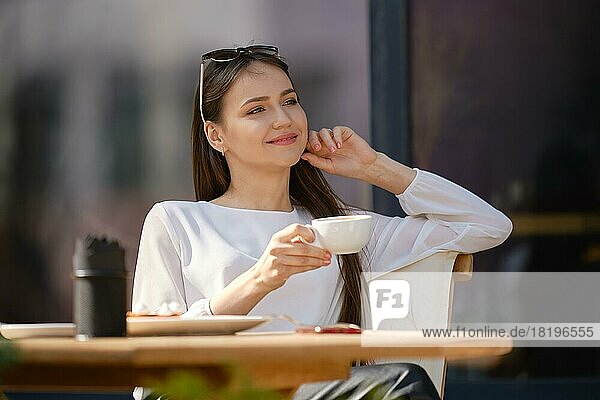 Verträumte junge Frau sitzt auf der Terrasse des Cafés und trinkt Cappuccino