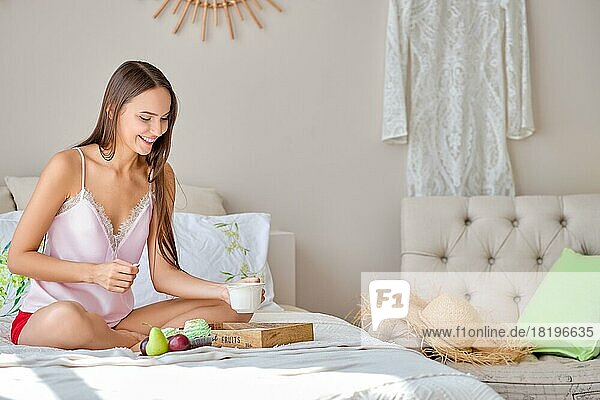Glückliche junge Frau  die eine Tasse Tee trinkt  während sie im Bett isst