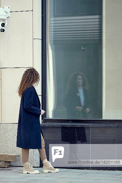 Unbekannte Frau im Mantel geht den Bürgersteig entlang und betrachtet ihr Spiegelbild in den Fenstern