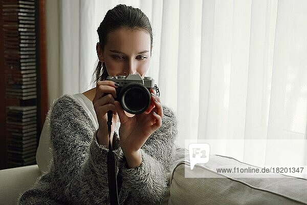 Glückliches lächelndes Mädchen mit Fotokamera und Smartphone in den Händen auf dem Sofa in der Nähe des Fensters