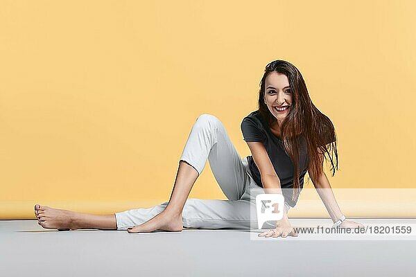 Glückliche barfüßige junge Frau in Freizeithose und Hemd sitzt auf dem Boden im Studio auf gelbem Hintergrund