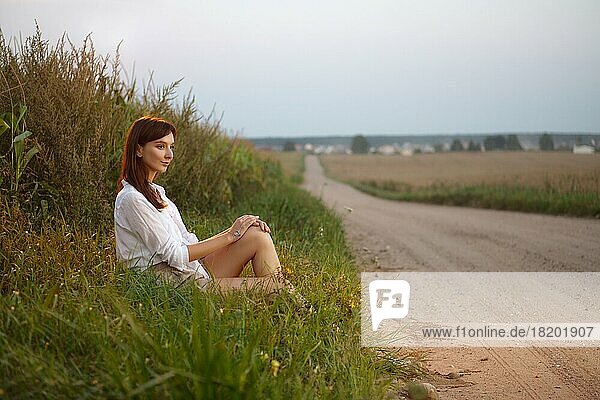 Hübsches Mädchen sitzt im Profil am Rande der Landstraße bei Sonnenuntergang