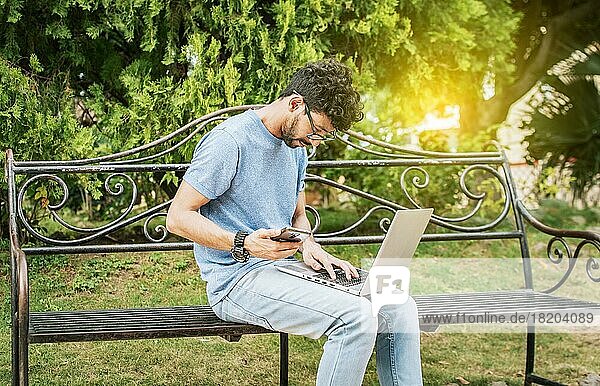 Young man in a park with laptop and cell phone. Freelancer man sitting in a park using laptop and cellphone. Man in a park working online with laptop. Relaxed man working with laptop outdoor
