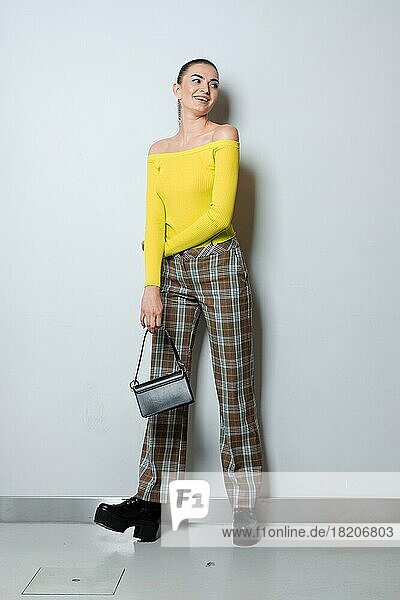Girl in plaid pants and yellow pullover with small handbag posing near white wall