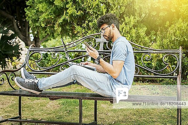 Man in a park working online with laptop. Relaxed man working with laptop outdoor  Young man in a park with laptop and cell phone. Freelancer man sitting in a park using laptop and cellphone