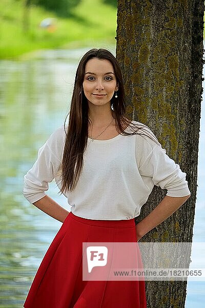 Portrait of a girl in red skirt and white blouse leaning against a tree on river bank