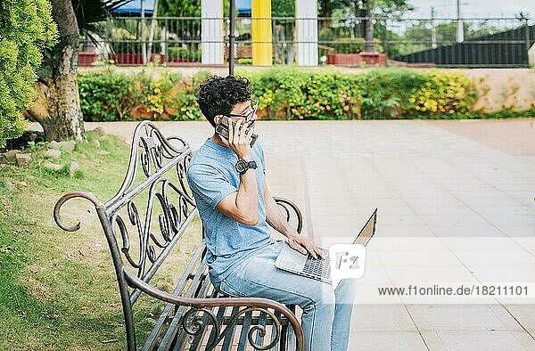 Junger Mann sitzt in einem Park und arbeitet mit seinem Laptop und seinem Mobiltelefon. Mann in einem Park arbeiten online mit Laptop und Anruf auf Handy. Freelancer Mann arbeitet mit Laptop und ruft am Telefon