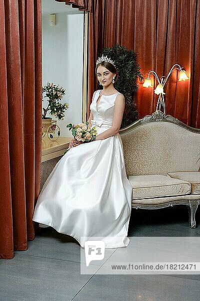 Beautiful bride in lond dress sits on the couch by the window