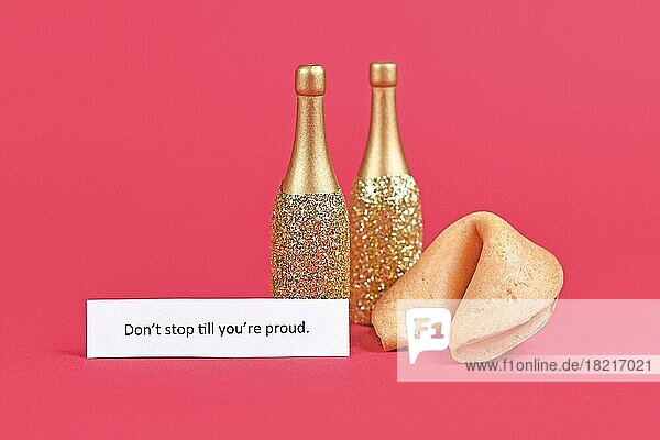 Glückskeks und Champagnerflaschen mit motivierendem Text 'Don't stop till you're proud' auf Papier auf rosa Hintergrund