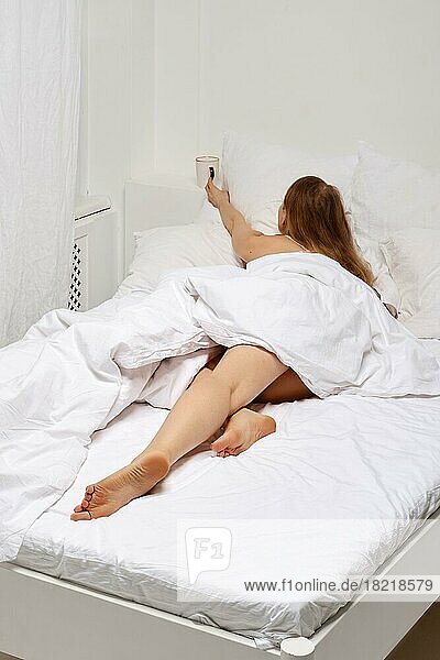 Unbekannte Frau liegt im Bett und greift mit der Hand zu einer Tasse Kaffee