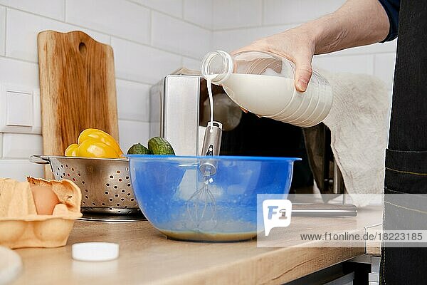 Nahaufnahme der Hände eines Mannes  der Milch in eine Schüssel mit geschlagenen Eiern gießt  um ein Omelett zu machen