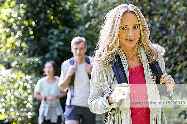 Mid-shot of blonde middle-age woman with backback hiking with her diverse group of friends in the woods