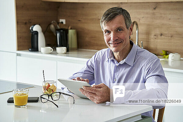 Middle-aged professional man looking at camera and checking messages on digital tablet while having breakfast on kitchen counter at home