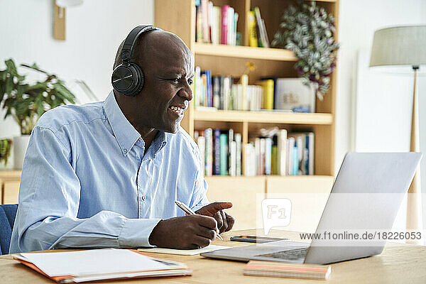 Middle-aged African-American man wearing headphones and taking notes while attending remote conference call while working at home with laptop computer