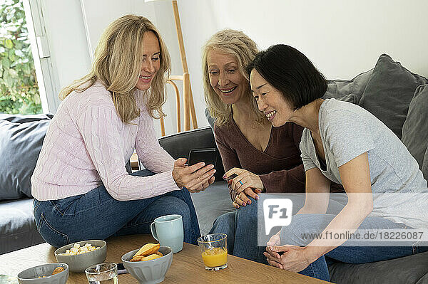 Portrait of three female middle-aged women having fun while looking at photos on phone in living room