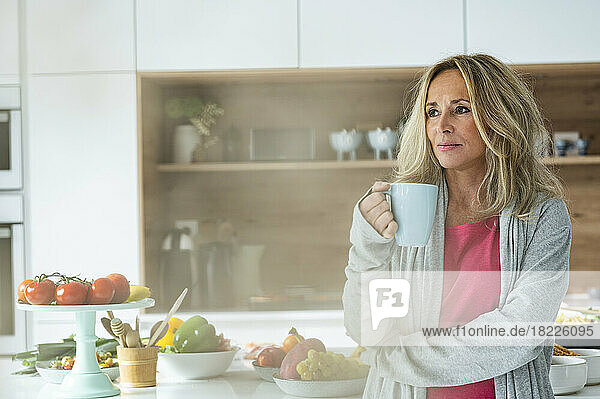 Portrait of attractive middle-aged woman holding a cup of coffee looking at camera standing in kitchen