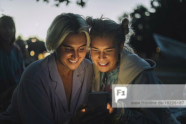 Glückliche nicht-binäre Person  die ihr Smartphone mit einer Freundin bei Sonnenuntergang teilt