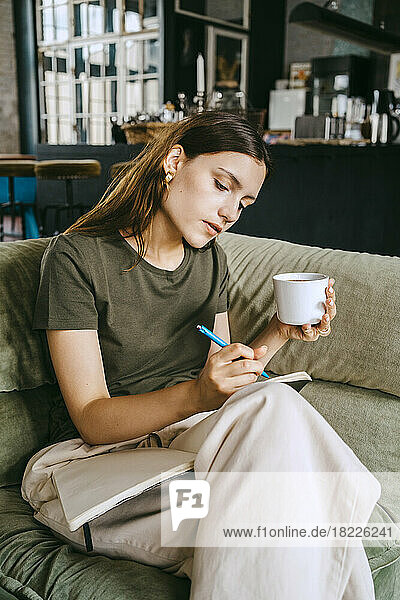 Junge Unternehmerin  die in ein Buch schreibt und eine Kaffeetasse hält  während sie auf dem Sofa im Studio sitzt