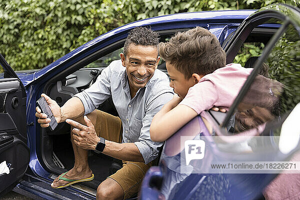 Excited father showing mobile phone to son leaning on car door