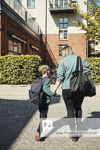 Rückansicht einer Mutter  die an einem sonnigen Tag mit ihrem Sohn in Richtung Schulgebäude geht