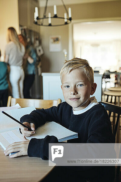 Porträt eines lächelnden Jungen  der mit einem Arbeitsbuch auf einem Tisch zu Hause sitzt