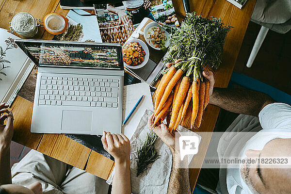 Ein männlicher Kollege hält ein Bündel Karotten in der Hand  während ein Unternehmer einen Laptop auf dem Tisch hat.