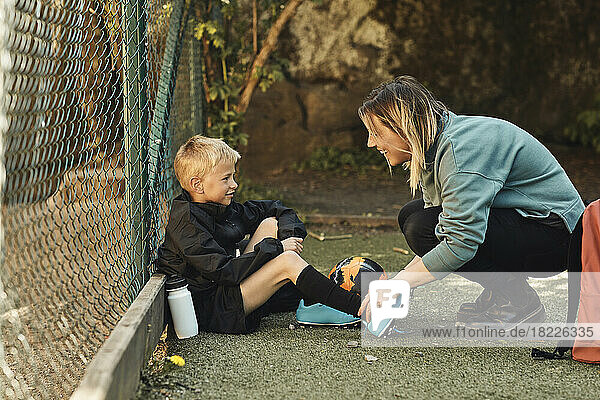 Happy woman tying shoelace of son sitting by fence in ground