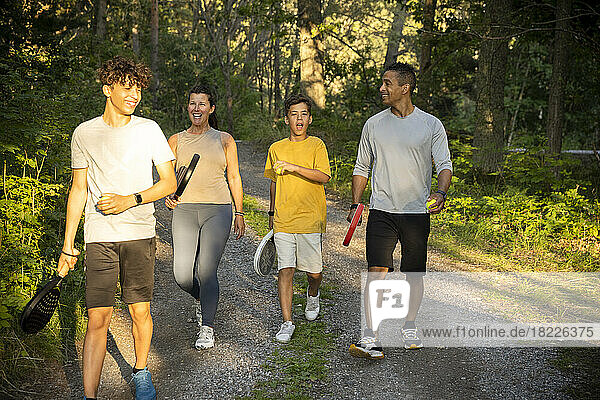 Glückliche Familie mit Tennisball und Schlägern  die im Urlaub auf einem unbefestigten Waldweg spazieren geht