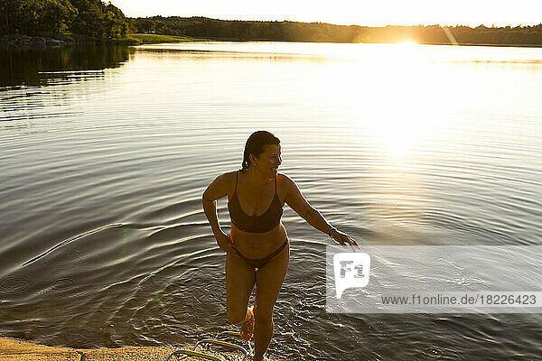 Happy woman in bikini walking out of lake after swimming during sunset